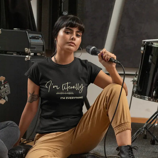 A woman sits on a wooden floor in a music studio, holding a microphone. She is wearing a black graphic t-shirt with the text 'I'm totally ANIMATED I'M EVERYTHING' in white script, paired with tan cargo pants and black lace-up boots.