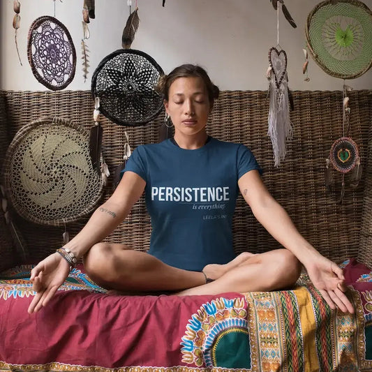 A woman sits cross-legged in a meditative pose on a woven bench, wearing a navy blue t-shirt with the text 'PERSISTENCE is everything' in white. The background features a wall decorated with multiple dreamcatchers, and the bench is draped with a red and patterned textile.