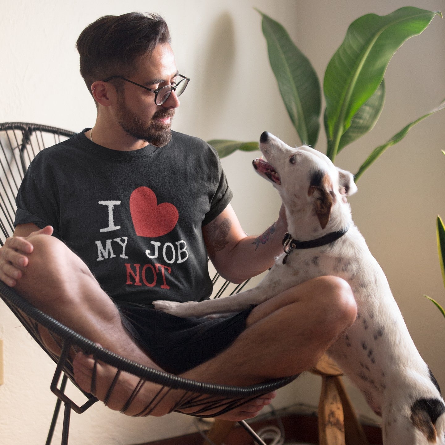 A man wearing a black graphic t-shirt with the text 'I [red heart] MY JOB NOT' sits cross-legged in a black wire chair, interacting with a white and black spotted dog in a home setting.