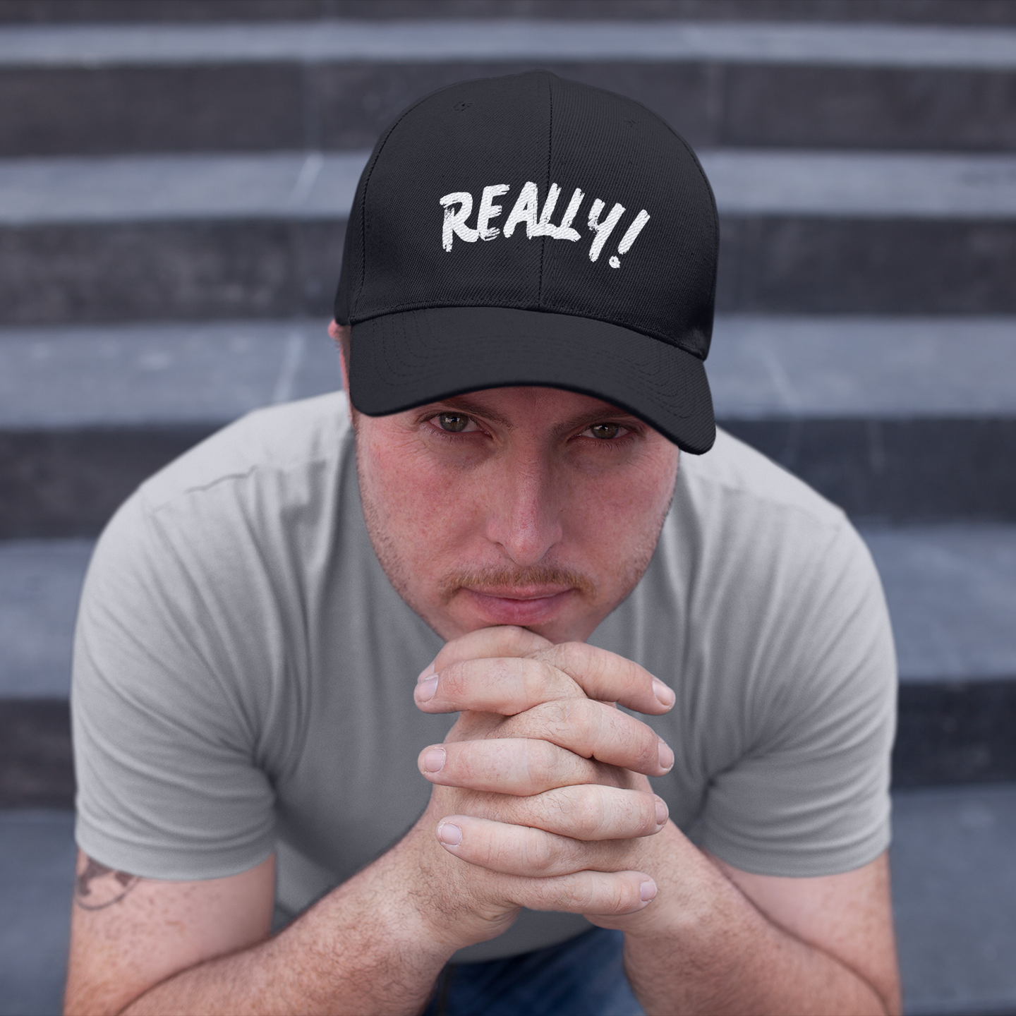 Really! embroidered black dad cap — worn by male model seated on steps, hands clasped